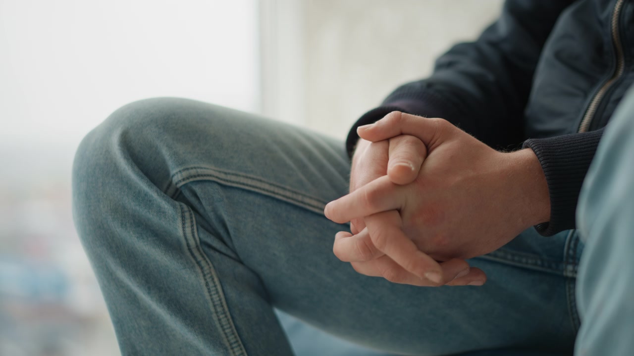 Primo piano delle mani di una persona giunte, appoggiate sulle ginocchia, che indossa jeans blu e una giacca scura. La scena è delicatamente illuminata da luce naturale proveniente da una finestra sullo sfondo, creando un'atmosfera interna calma.