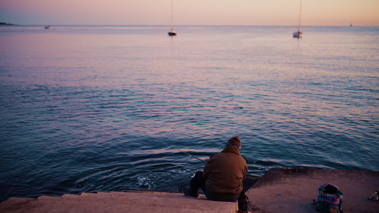 A man sits at the water's edge holding a fishing rod as the sky glows softly at sunrise
