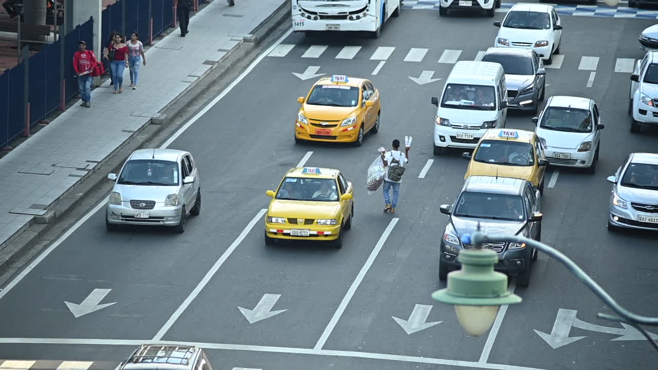 Cars stopping at red light in down town main avenue of Guayaquil (Malecon) and then moving forward. Some people passing by on sidewalk. A boy selling water bottles walks in the middle of the avenue.