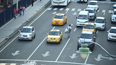 cars stopping at red light in down town main avenue of guayaquil (malecon) and then moving forward. some people passing by on sidewalk. a boy selling water bottles walks in the middle of the avenue.