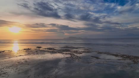 Panning-drone-clip-of-stunning-golden-sunset-reflecting-over-ocean-and-Indonesian-coral-reef-with-coastal-landscape-in-the-background