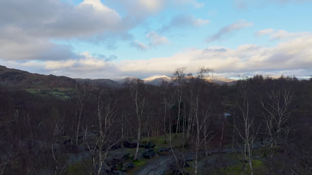 A drone lifts along a quiet path lined by bare winter trees, revealing distant mountains where sunlight breaks through clouds over the scenic landscape