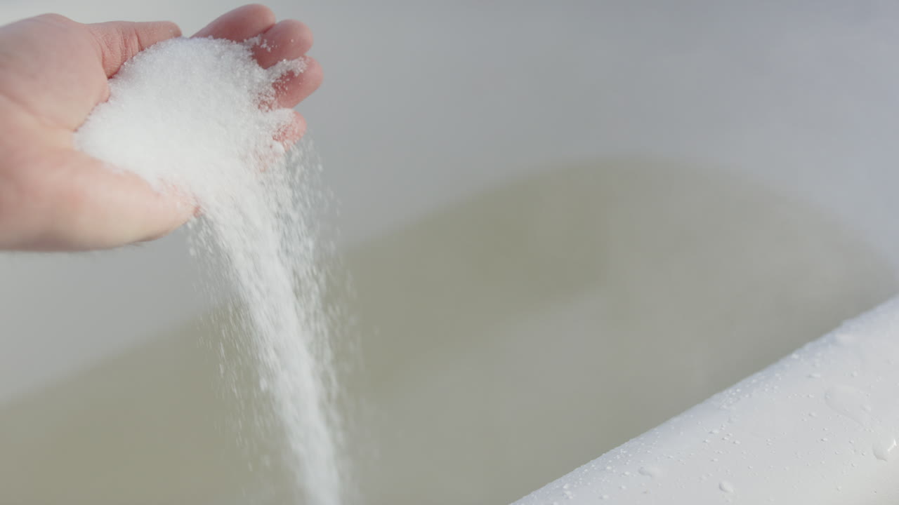 Epsom salts being dropped in slow motion from hand into steaming bath