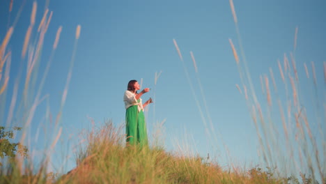 woman standing on grassy hilltop near cliff blowing delicate soap bubbles into clear blue sky, green dress fluttering in breeze as sunlight warms landscape and bubbles catch rainbow reflections