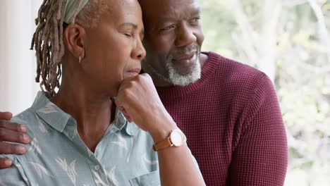 Nachdenkliches-Afroamerikanisches-Seniorenpaar,-Das-Sich-Zu-Hause-Umarmt-Und-Aus-Dem-Fenster-Schaut,-Zeitlupe