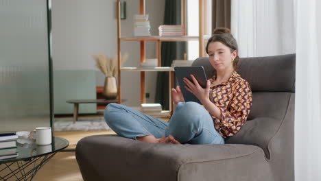 Young-woman-relaxes-and-uses-tablet-in-modern-living-room,-full-shot