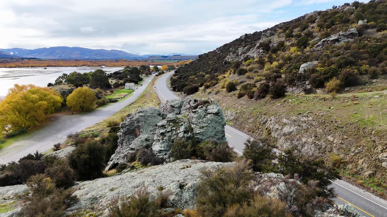 Drone glides above a winding lakeside road bordered by rocky outcrops, autumn trees, and distant mountains under overcast daylight, revealing scenic natural beauty