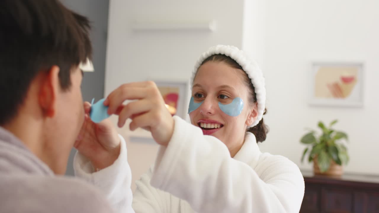 Couple in white robes on bed, woman offering blue under-eye patch and placing it for skincare