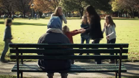 a solitary figure sits on a park bench, observing as a group of friends joyfully plays and dances in the vibrant autumn landscape, surrounded by colorful leaves and sunshine.