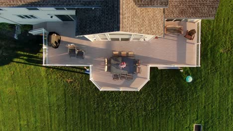 rising aerial top down view of large deck, outdoor living space with white railing, table, chairs, grill among green grass yard