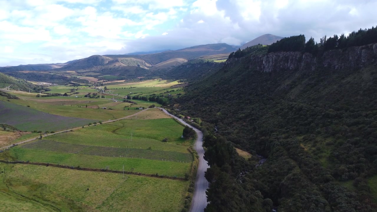 Vista aerea con drone di campi coltivati nella valle nelle Ande ecuadoriane, incorniciati da una lunga scogliera rocciosa, con una strada sterrata che conduce verso distanti montagne nuvolose