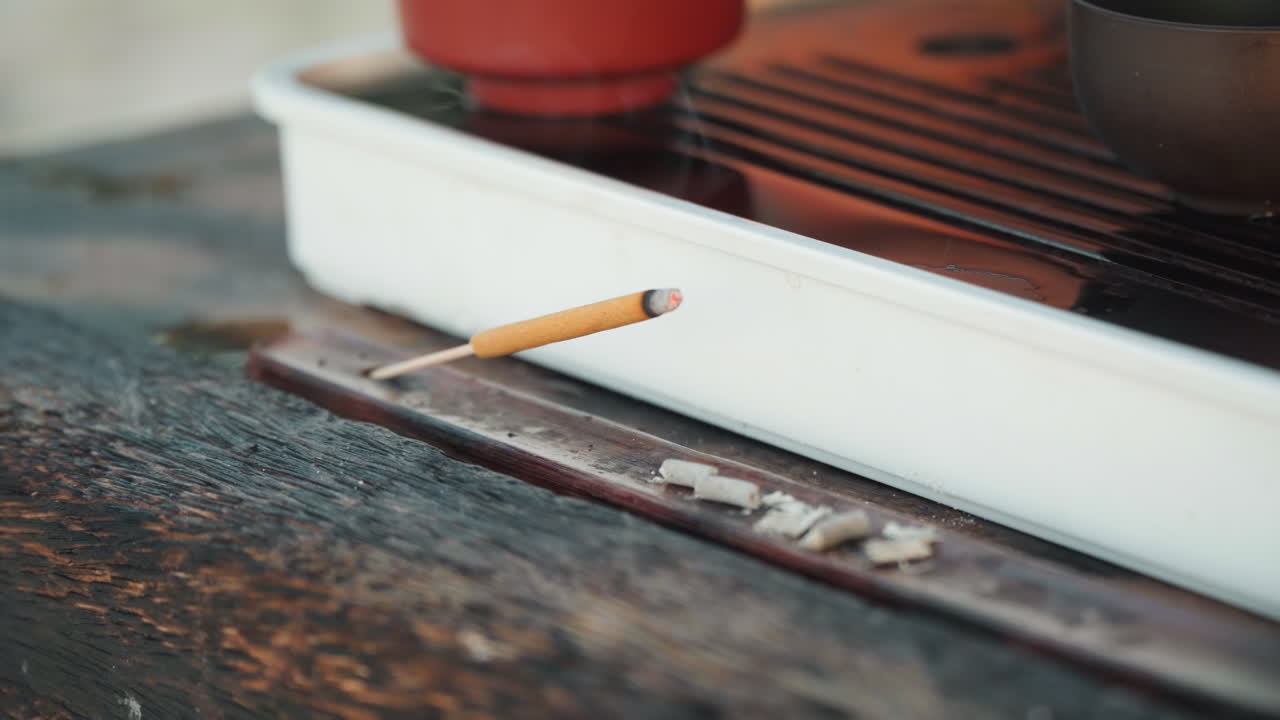 陶器の茶盆の横にあるマッチのクローズアップ。風化した木材の表面で行われる海辺の儀式。落ち着いた色調、消えた残り火から穏やかに立ち上る煙、温かい琥珀色のトーン。マインドフルなスローライフの雰囲気。