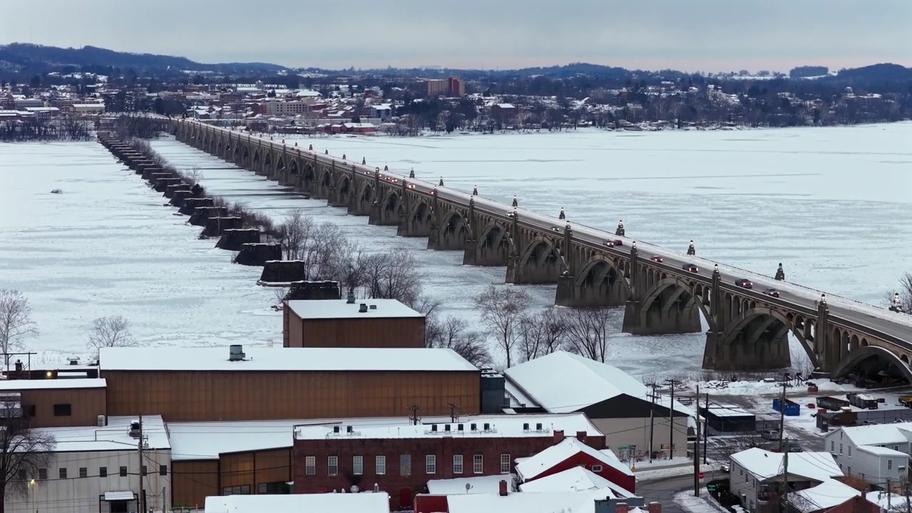 Città di Wrightsville, Pennsylvania, coperta di neve con ponte storico che attraversa un fiume ghiacciato, traffico invernale e tetti sotto un cielo freddo e coperto. Atmosfera stagionale nevosa e cinematografica. Ampia ripresa aerea.