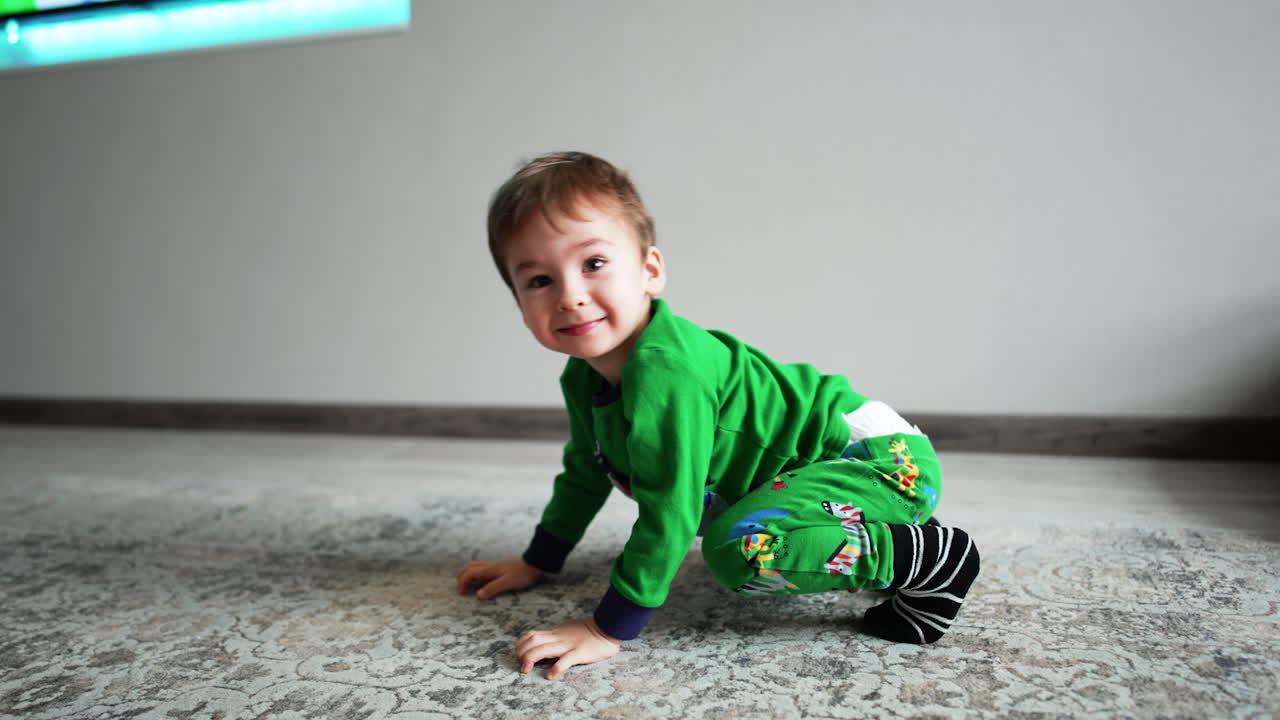Lovely toddler male kid in green pajama tries to stand on his arms. Little child tumbles on the floor.