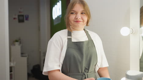 beautician in white shirt and gray apron walking toward client with warm smile while holding blue cup and saucer, offering tea in cozy salon with soft lighting and modern interior decoration