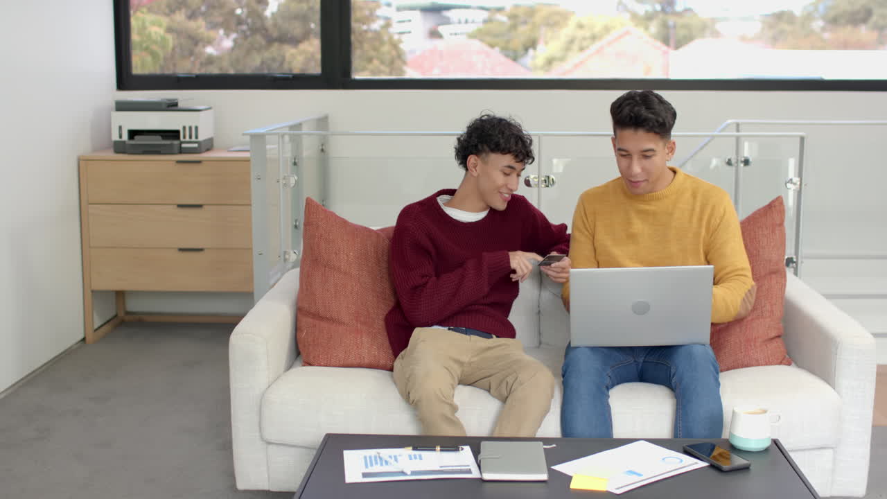 Sitting on couch, multiracial gay couple using laptop and credit card for online shopping, at home
