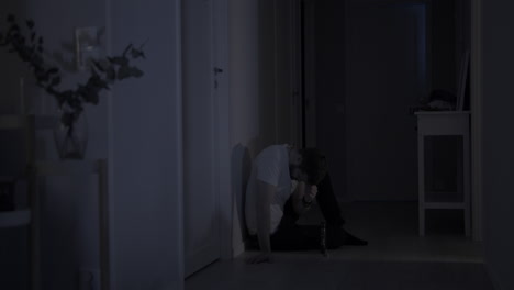 Depressed-Man-in-Grief-sits-alone-on-Home-Floor-leaning-against-dark-wall