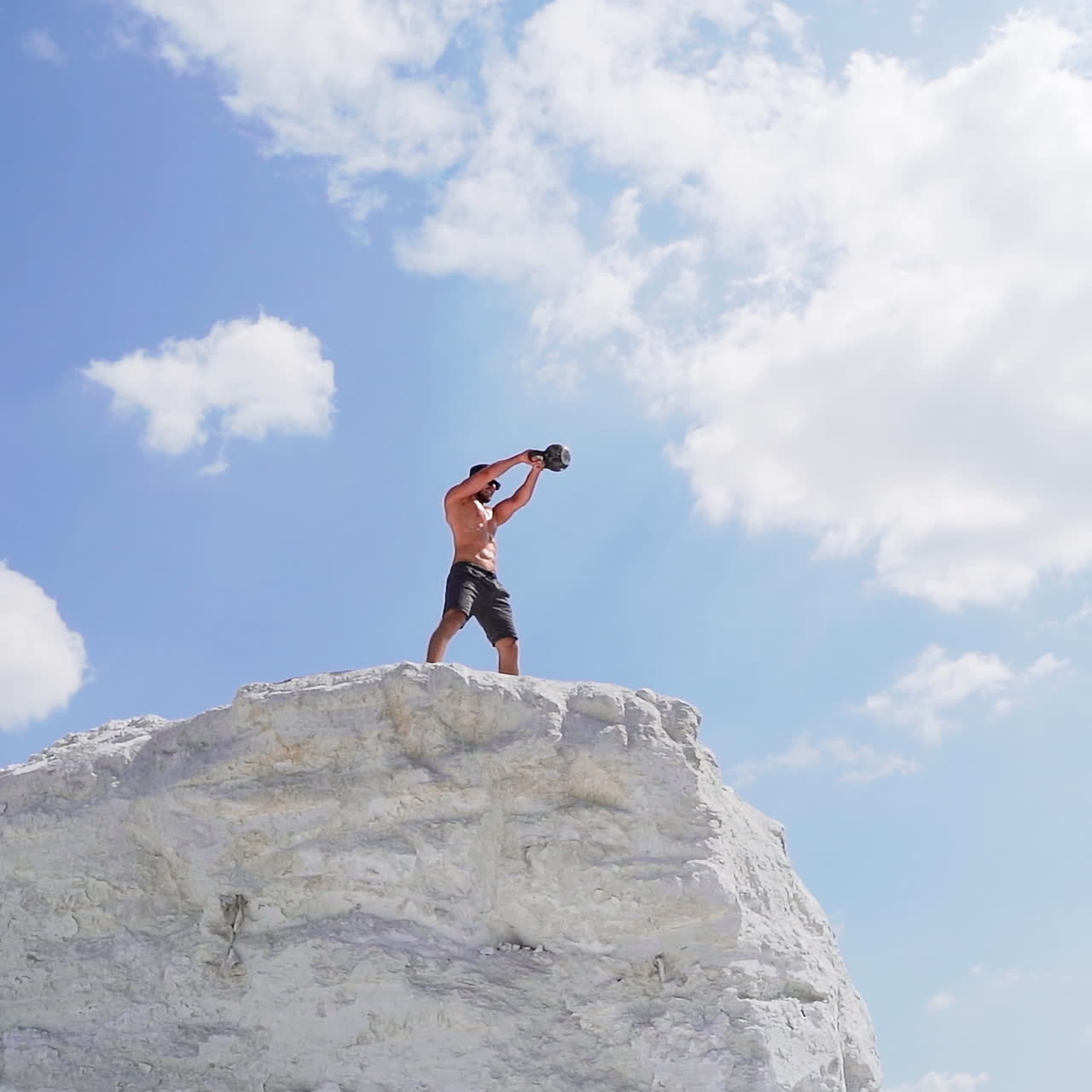 Shirtless athlete lifting kettlebell. Healthy sportsman is swinging kettlebell with two hands on the top of white hill. View from below on bodybuilder under blue sky.