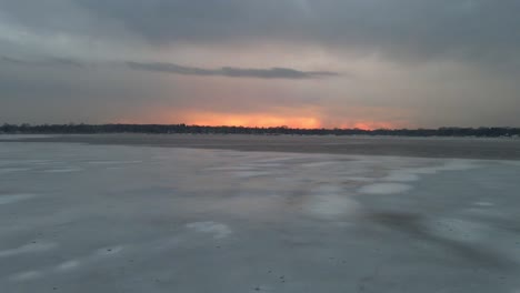 Vista-Aérea-De-Un-Lago-Congelado-Con-Patrones-Geniales-En-El-Hielo-Con-Una-Colorida-Puesta-De-Sol-En-El-Horizonte