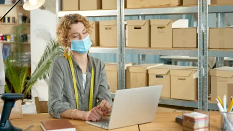 young caucasian designer woman in facial mask sitting at desk and videochatting on laptop in clothing shop warehouse