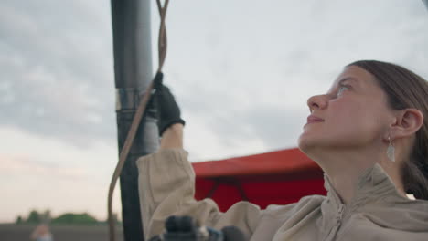 close up of focused woman in beige jacket loosening rope while gazing upward near hot air balloon basket during soft sunset light, calm sky and subtle tension