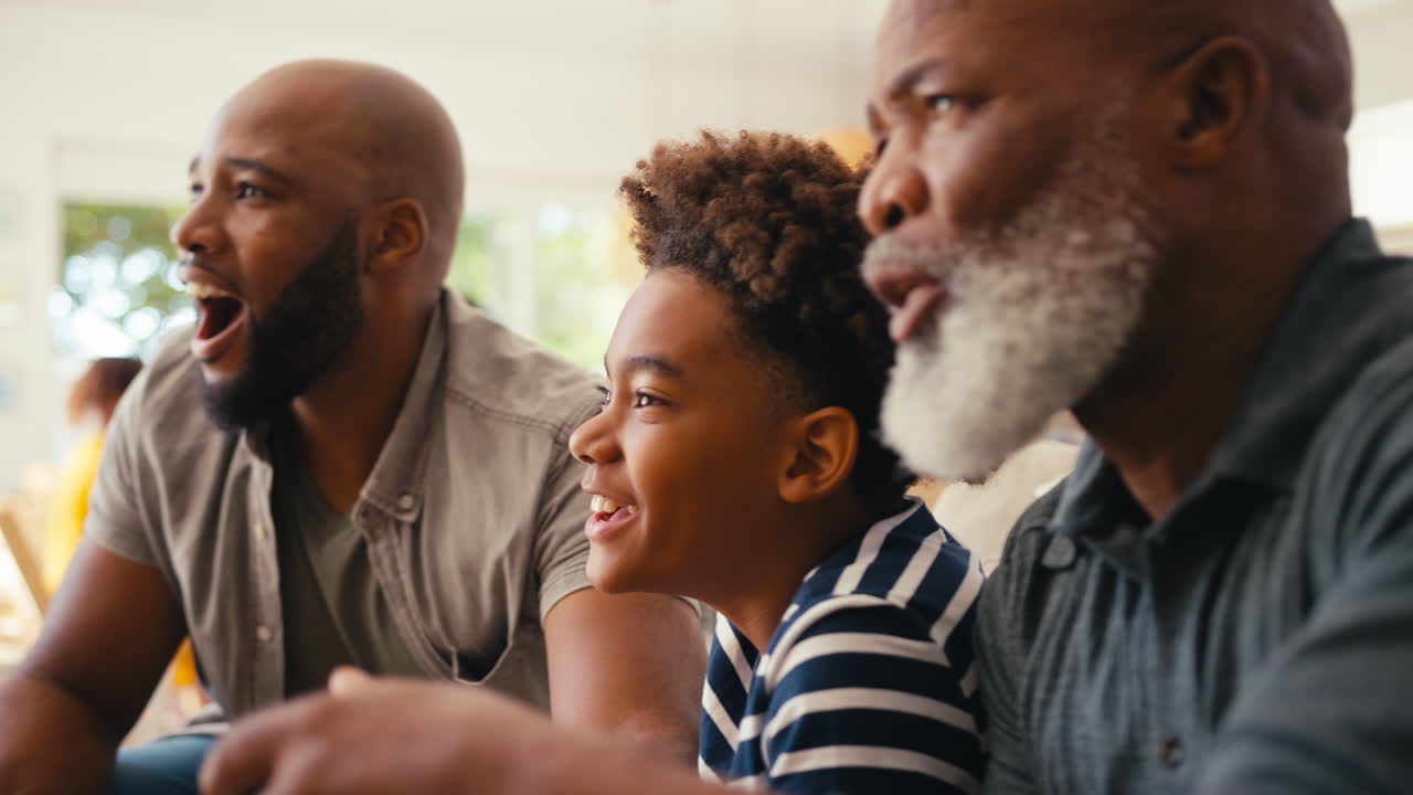 Close Up Of Multi-Generation Male Family Sitting On Sofa At Home Playing Video Game Together