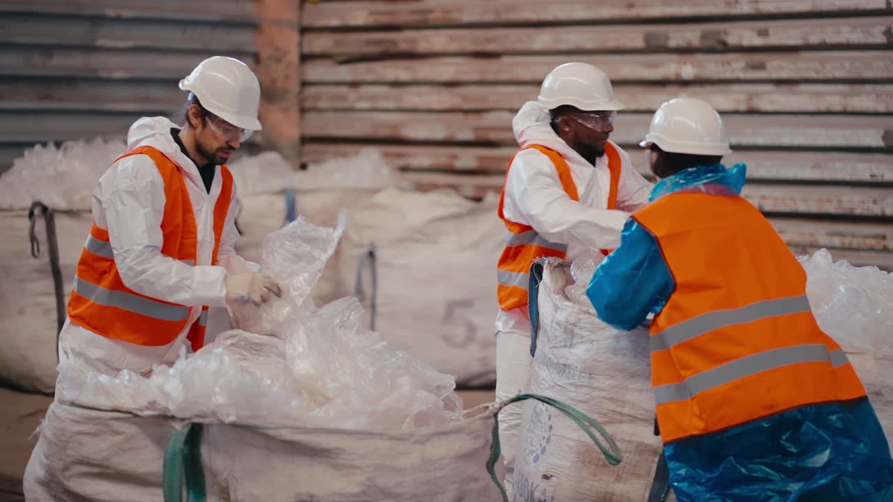 un equipo de tres trabajadores en un uniforme blanco y un chaleco naranja apilan y prensan polietileno y celofán mientras trabajan en una gran planta de reciclaje de residuos