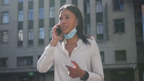 mujer afroamericana con máscara facial hablando en un teléfono inteligente en la calle de la ciudad