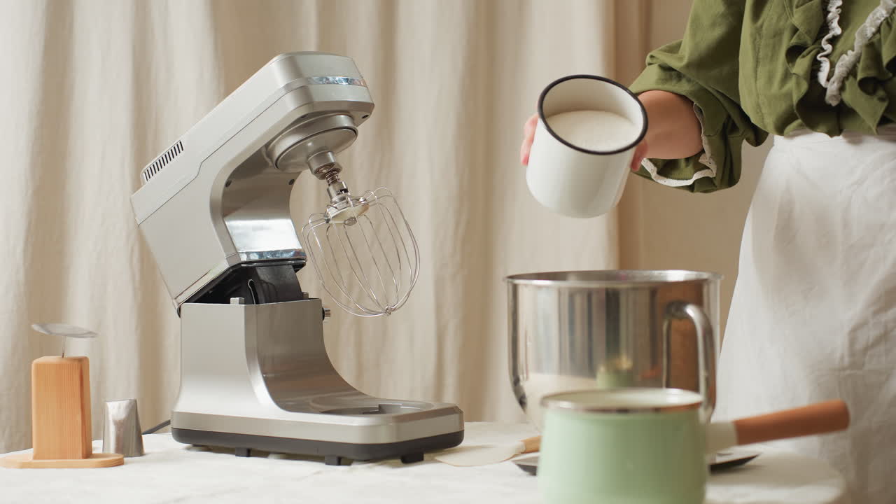 Chef wearing green top and white apron preparing ingredients in kitchen by pouring sugar from white metal cup into stainless mixing bowl beside electric stand mixer on countertop