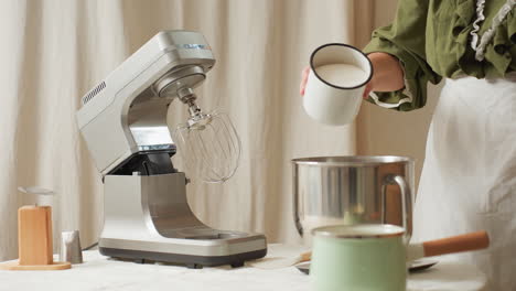 chef wearing green top and white apron preparing ingredients in kitchen by pouring sugar from white metal cup into stainless mixing bowl beside electric stand mixer on countertop