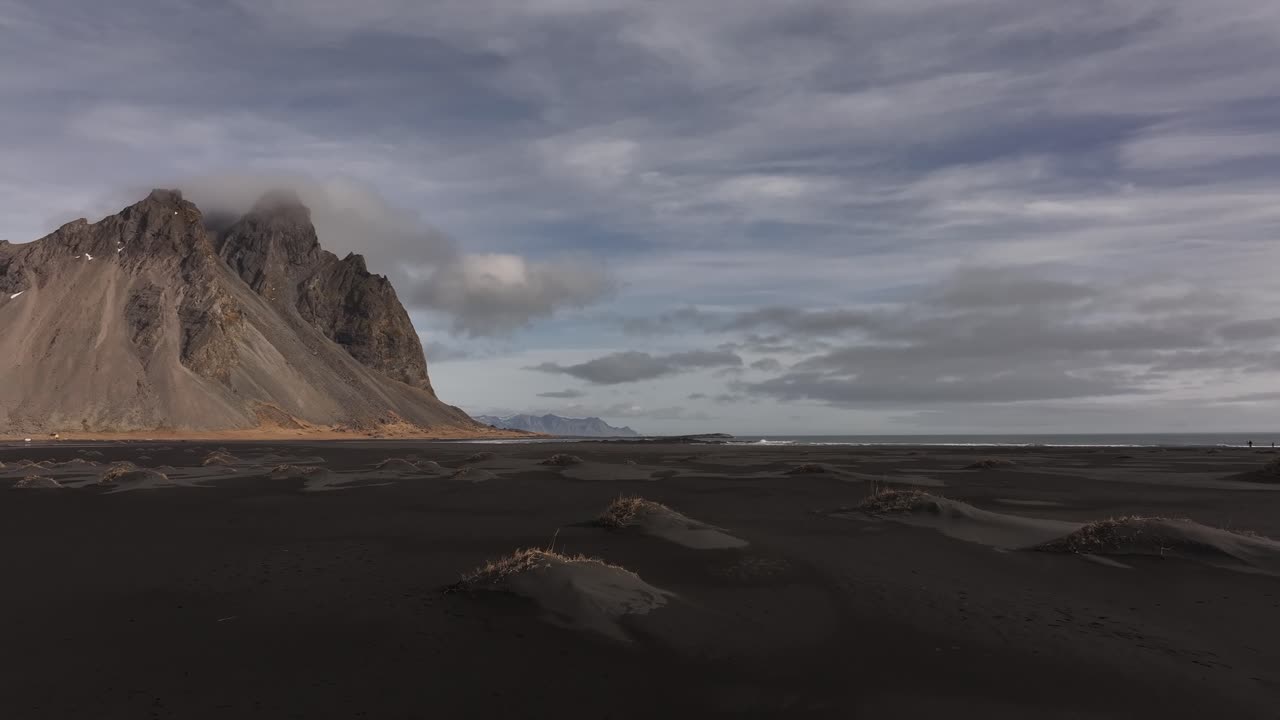 Drone moving forward over a black sand dune field, approaching the dramatic Horny Mountains in Stokksnes, Iceland, under a moody sky.