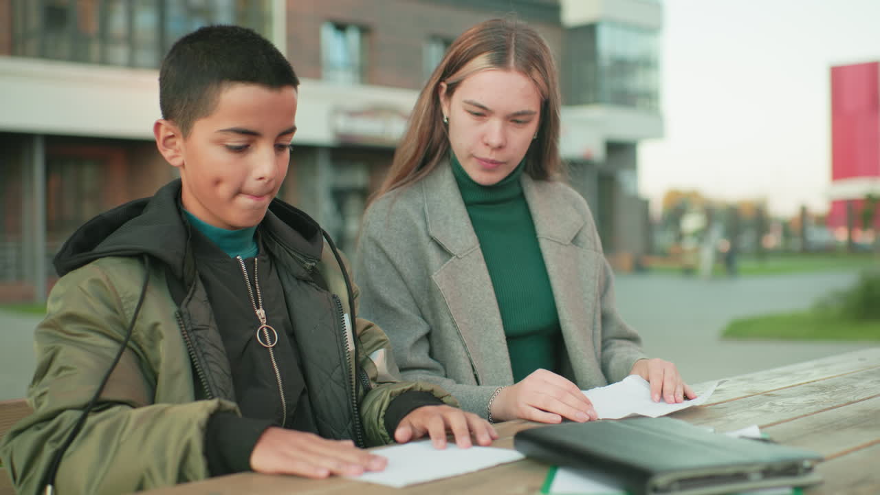 Lady shows young boy how to make paper craft while seated at outdoor wooden table, both focused on folding sheets, emphasizing creativity, family connection