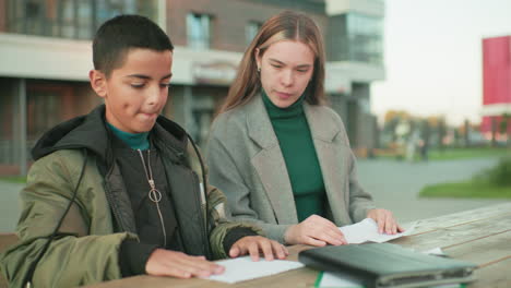 lady shows young boy how to make paper craft while seated at outdoor wooden table, both focused on folding sheets, emphasizing creativity, family connection