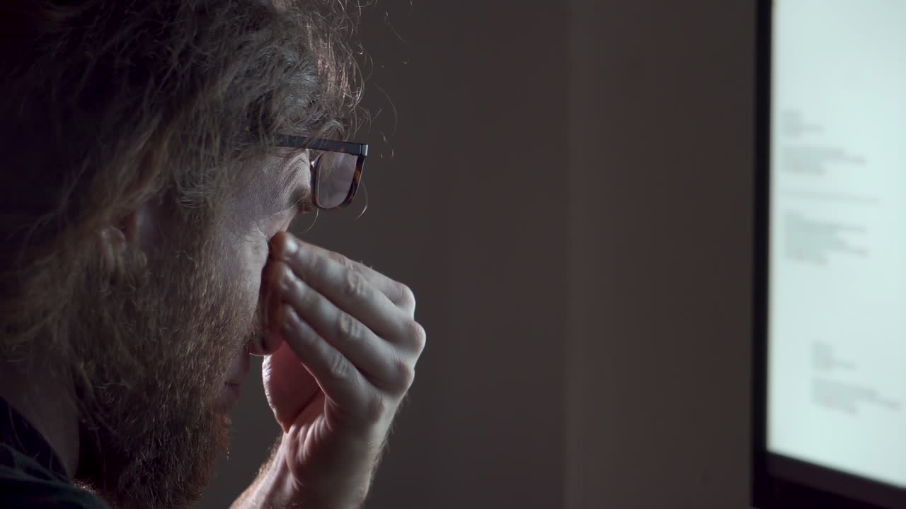 un hombre barbudo cansado que trabaja tarde en la noche en una oficina con poca luz se frota los ojos y la nariz y está exhausto