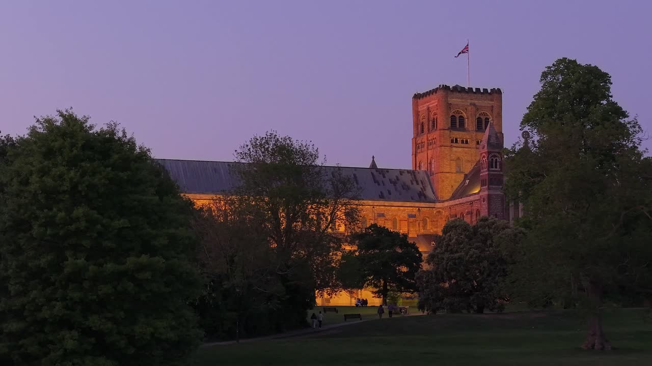 Static aerial or ground shot captures the illuminated St Albans Cathedral at night, with pedestrians walking along the park path, highlighting warm lights and evening atmosphere