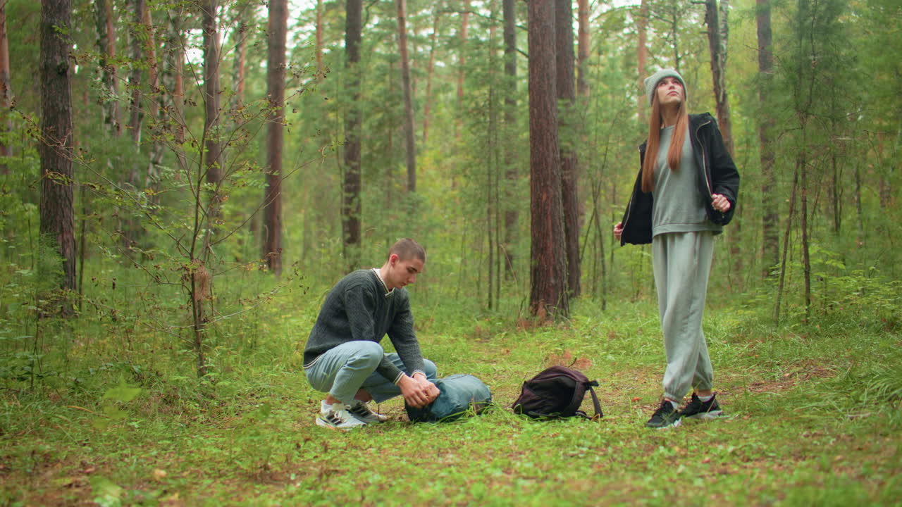 researchers in forest as man crouches to open green backpack on ground while woman stands observing woodland, dressed in casual clothing, surrounded by tall trees and dense greenery