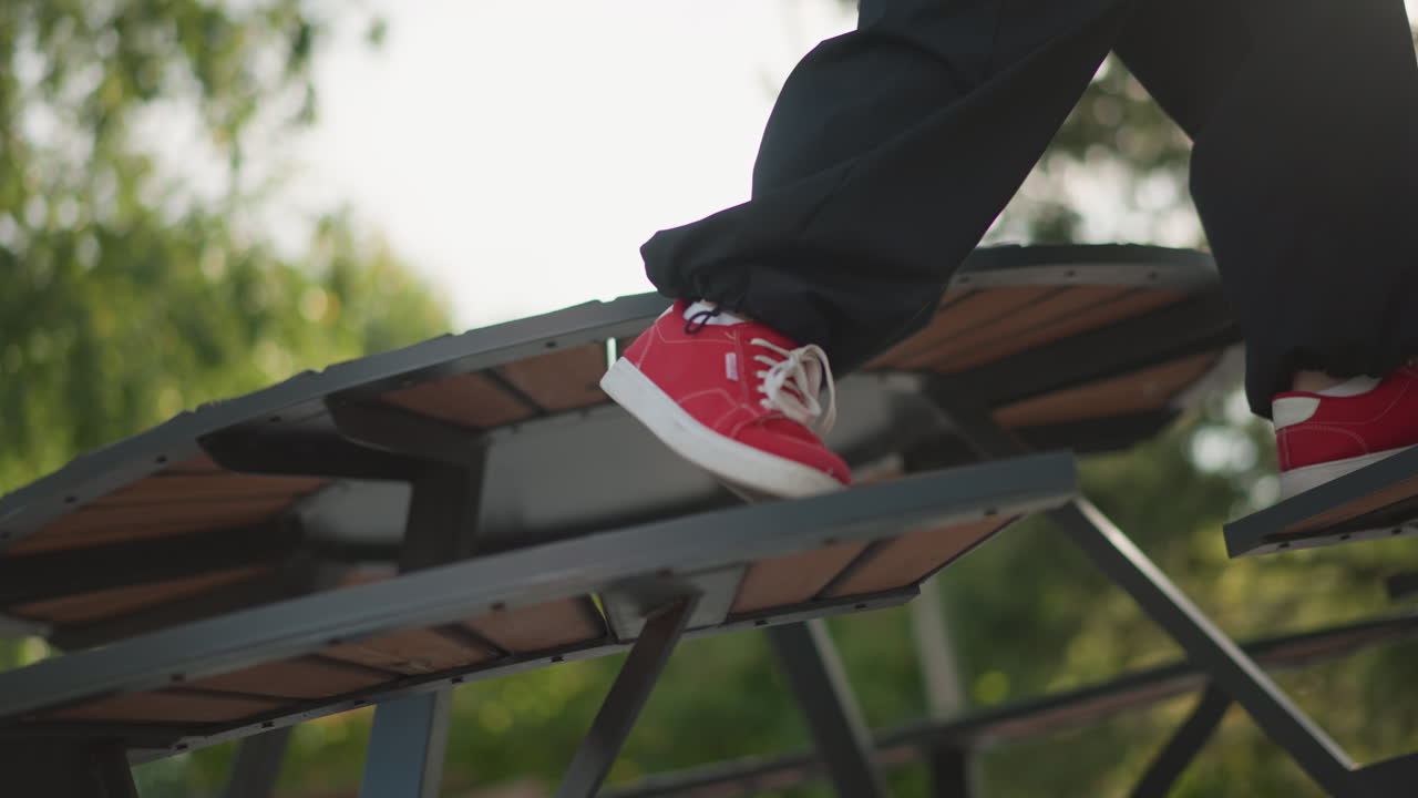 Gioco di gambe street dance su una panchina del parco che mostra sneakers rosse che si muovono su listelli di un tavolo in legno, primo piano delle gambe e pantaloni neri casual, equilibrio e movimento focalizzati, dettagli della struttura in metallo, tardo pomeriggio