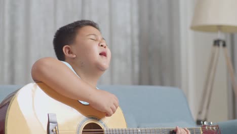 close up of talented asian kid with expressive face enjoys singing and playing the guitar on sofa at home