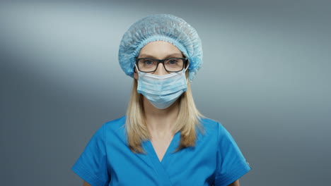 Portrait-Shot-Of-The-Young-Attractive-Blond-Female-Physician-In-The-Blue-Hat-And-Glasses-Taking-Off-Her-Mask-From-The-Mouth-And-Smiling-Happily