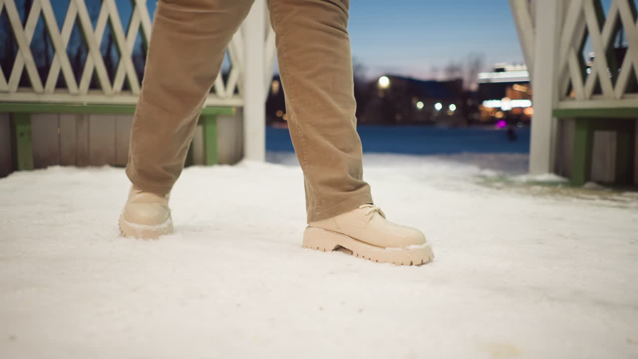 Woman wearing beige boots and khaki pants spins around shelter on snow covered floor under pavilion lights at dusk, viewed from low angle with rhythmic motion and soft ambient glow