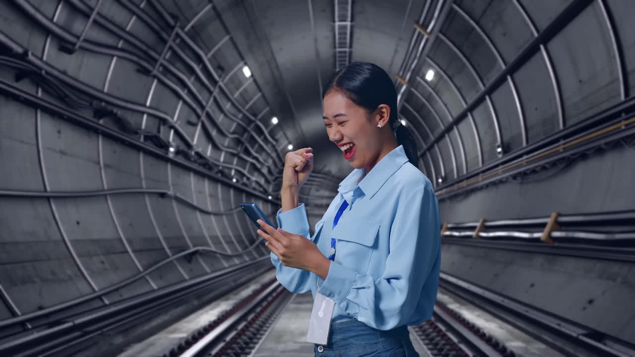 Side View Of Asian Female Professional Worker With Her Smartphone In Underground Subway Tunnel, She Raises Her Fist Up With Screaming Goal After Check On The Smartphone