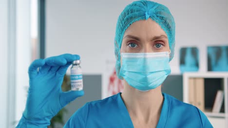 close up of female healthcare specialist virologist in protective uniform and medical mask holding in hand in gloves ampoule with coronavirus vaccine, covid 19 vaccination concept virus cure