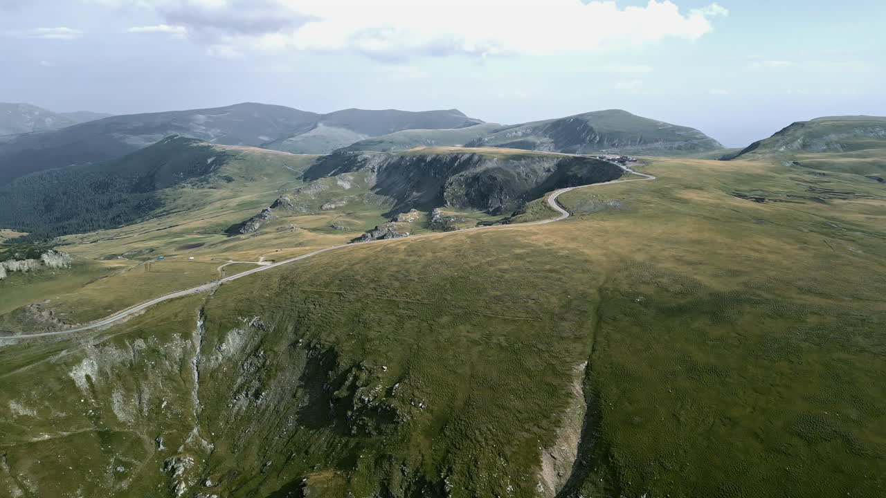 Elevated view of Romania's Transalpina, highlighting winding roads, verdant plains, and craggy cliffs set against a vast backdrop of rolling hills