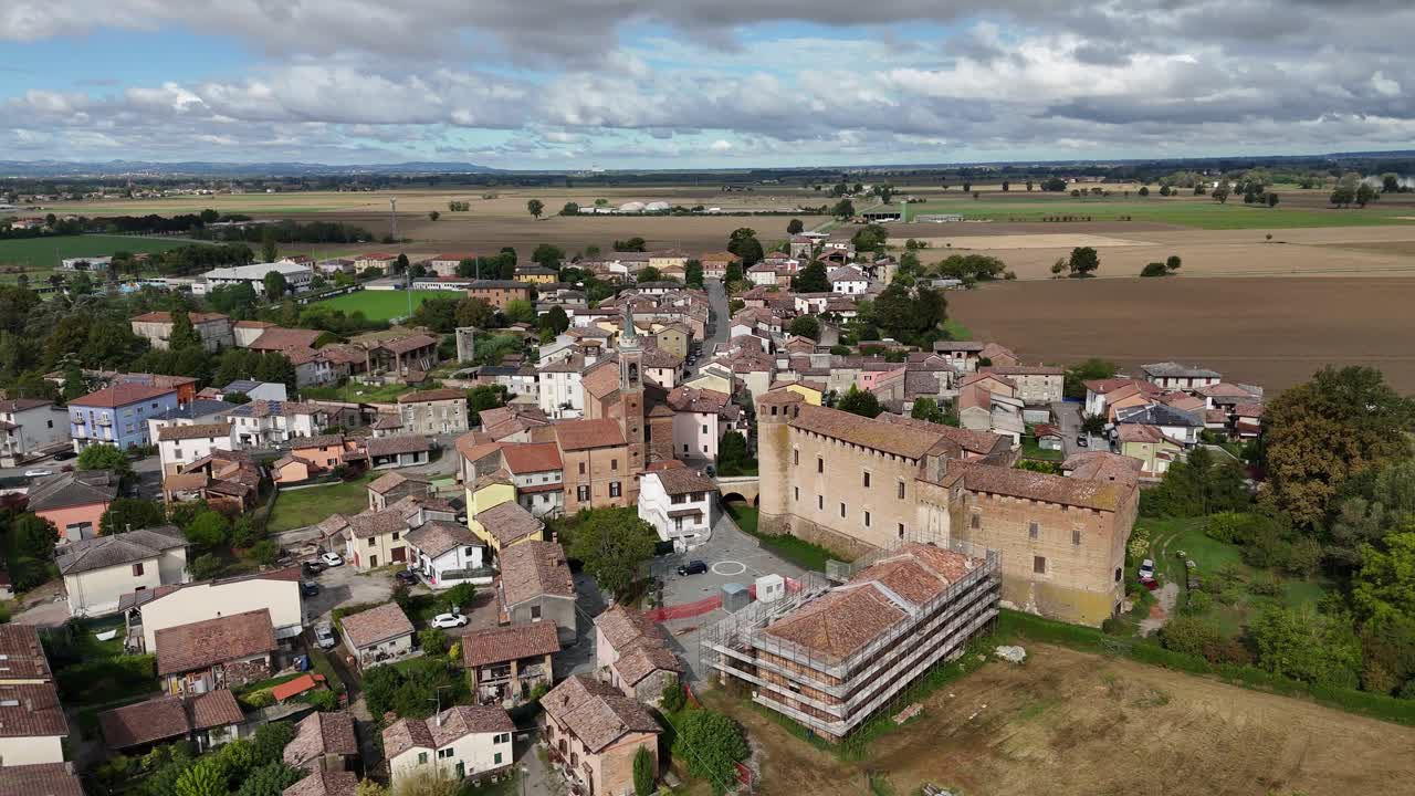 Campanile della chiesa parrocchiale di Calendasco e complesso del castello medievale a Calendasco, provincia di Piacenza, Italia, con tetti in terracotta, strade del borgo e terreni agricoli della Pianura Padana sotto nuvole pesanti, panoramica drone.