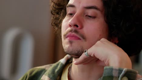 Close-up-of-a-sad-brunette-guy-with-curly-hair-holds-his-head-with-his-hand-and-thinks-hard-at-home-in-a-modern-apartment