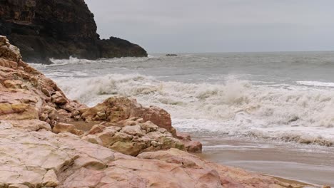 SLOW-MOTION-waves-white-with-foam-break-on-a-sandy-rocky-cliff-with-dark-stony-mountains-in-the-background,-Cape-Roca,-Portugal