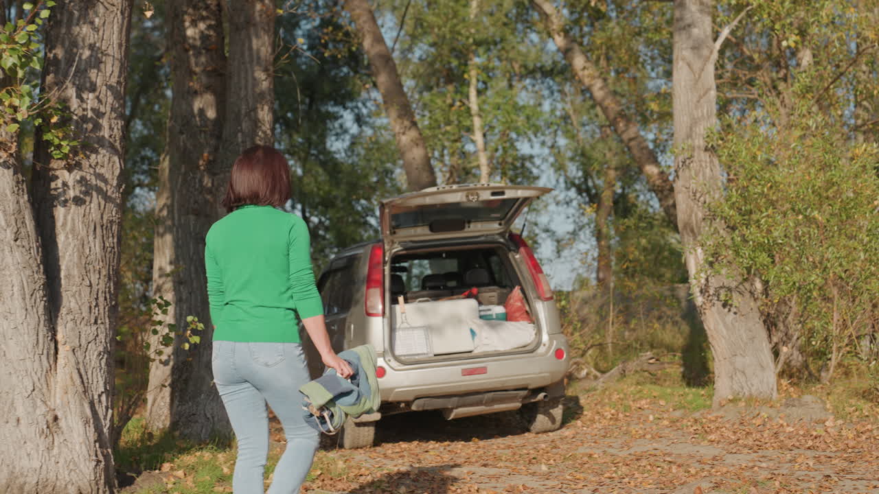 En hvit kvinne går mot en bil med åpent bagasjerom for å hente campingutstyr fra kombikupéen. Scenen er en høstskog med sollys som filtreres gjennom høye trær og løv under føttene. Hun er kledd i en uformell grønn genser og jeans.