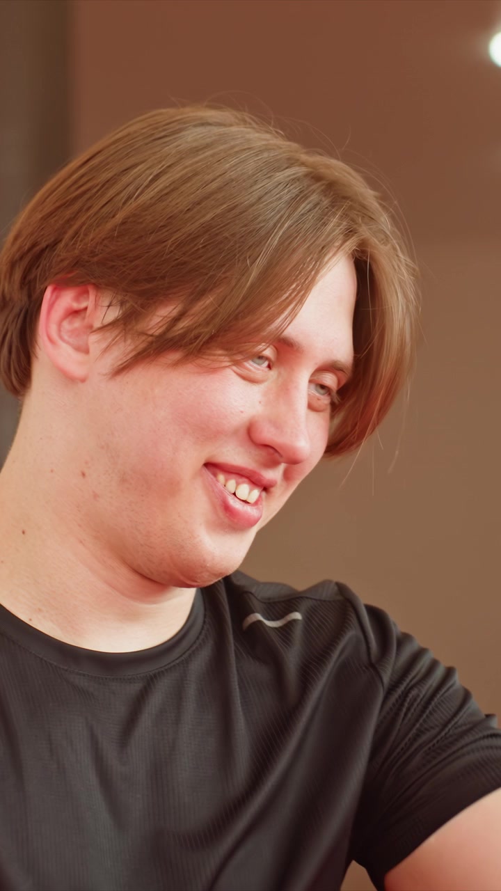 Close up of shirtless young man with tattoos smiling. Hand extends forward another man in black reacts. Warm lighting highlights skin tones against neutral beige walls