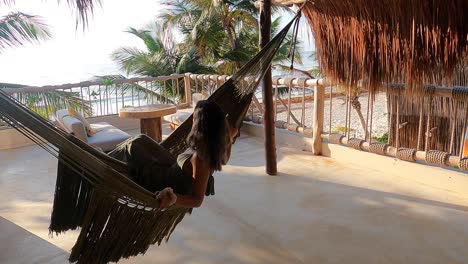 Drohnenaufnahme-Eines-Mädchens,-Das-Auf-Einer-Hängematte-Auf-Einem-Dach-Mit-Wunderschönem-Blick-Auf-Den-Strand-In-Tulum-Schwingt