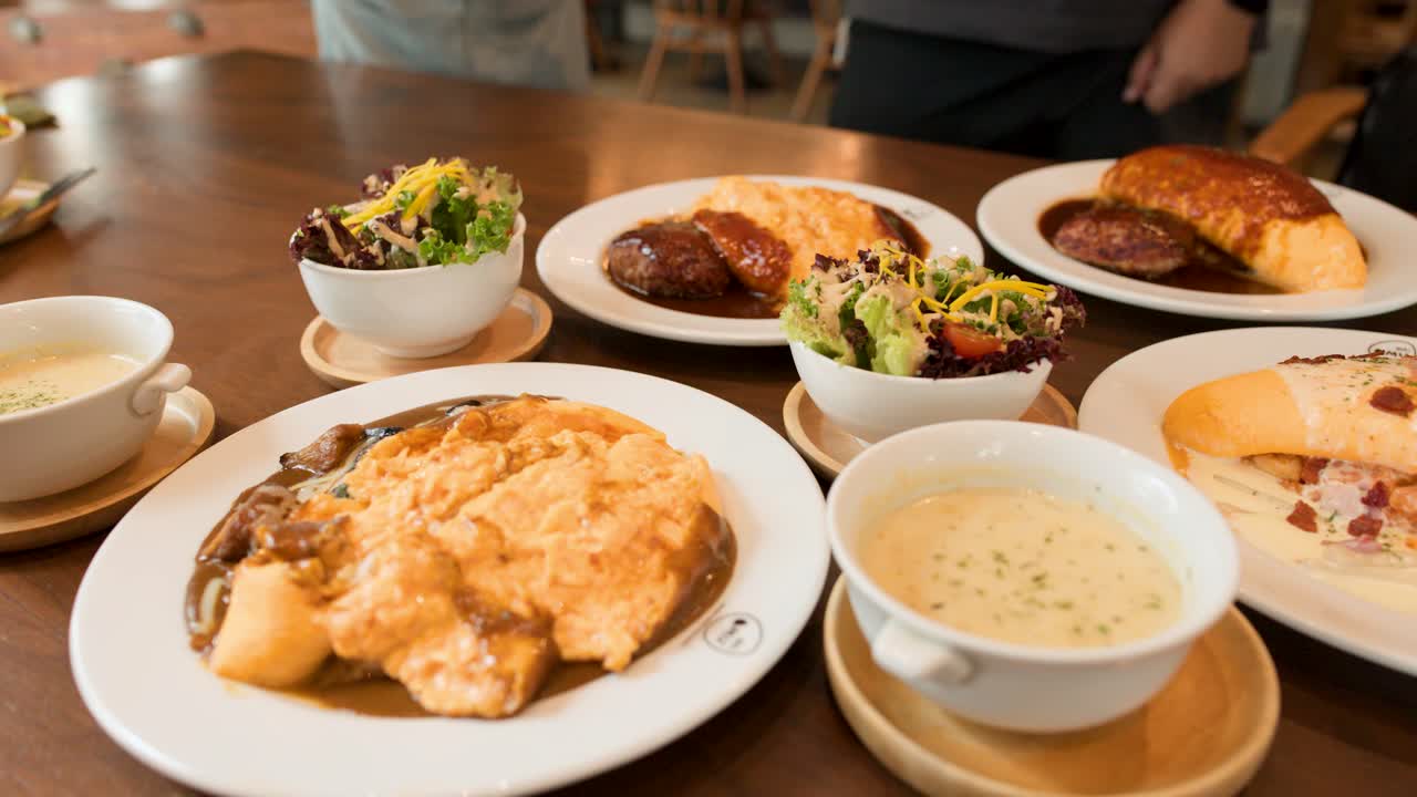 Hands serve Japanese omelette rice, creamy soup, salad, and bread in warmly lit restaurant